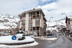 Um edifício com várias andares e varandas, uma rua coberta de neve e montanhas ao fundo.