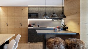 A modern kitchen with wooden cabinets, a dark countertop, hanging pendant lights, and a bar area with stools. - Pandora Mountain View - Central Apartment (Breuil-Cervinia)