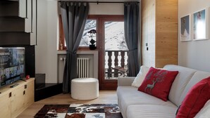 A living room with a television, a sofa, a round ottoman, and a view of a snowy landscape through a window. - Pandora Mountain View - Central Apartment (Breuil-Cervinia)