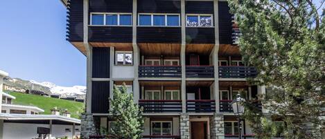 A multi-story building with balconies, a stone facade, and a view of snow-capped mountains.