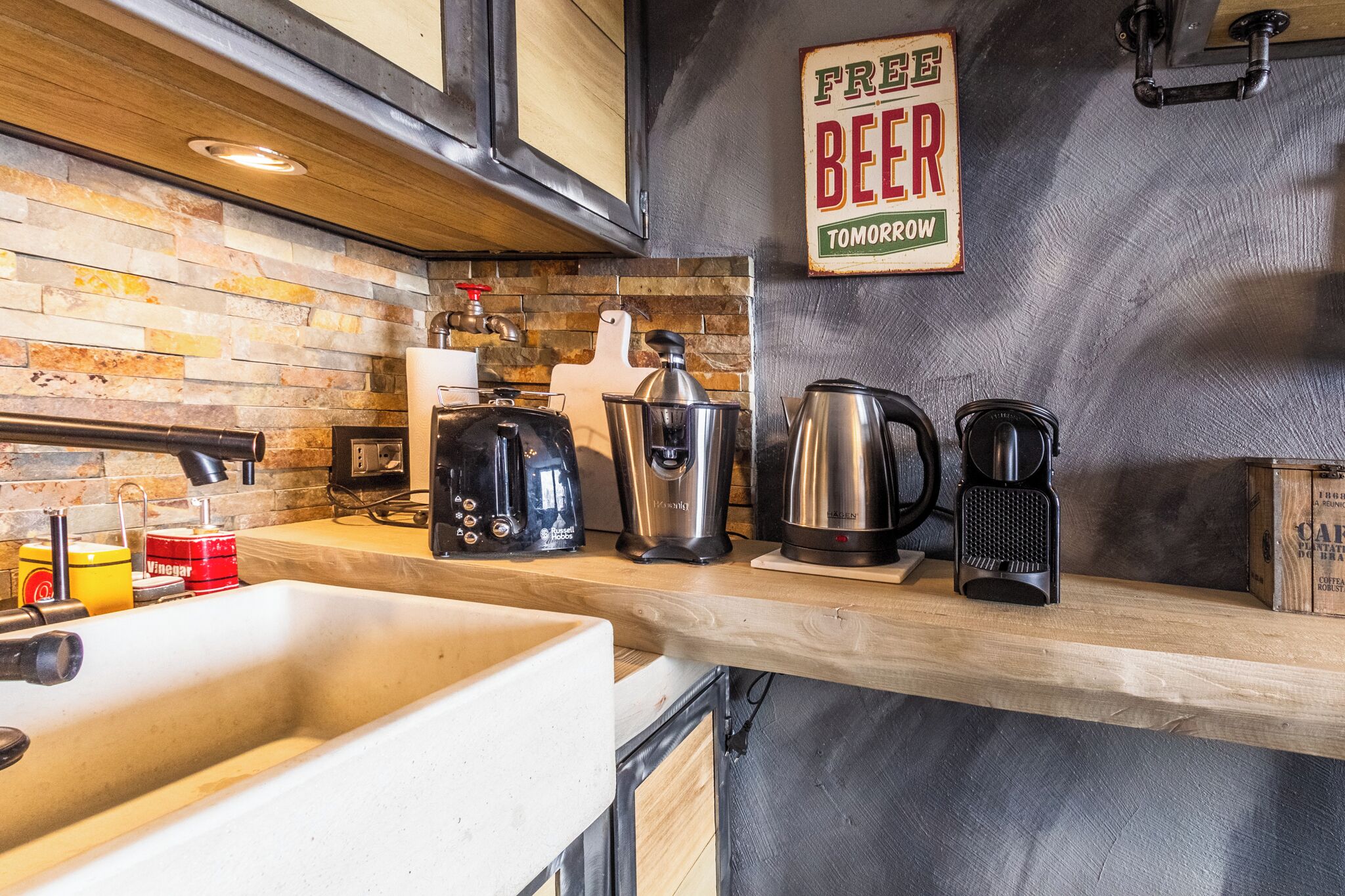 Een keuken met een achterwand van steen, een koffieapparaat, een waterkoker, en een bordje met de tekst 'GRATIS BIER MORGEN'.