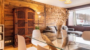 Una habitación con paneles de madera, una mesa de comedor de vidrio, sillas blancas, un sofá y un ventanal con vistas a un paisaje nevado.