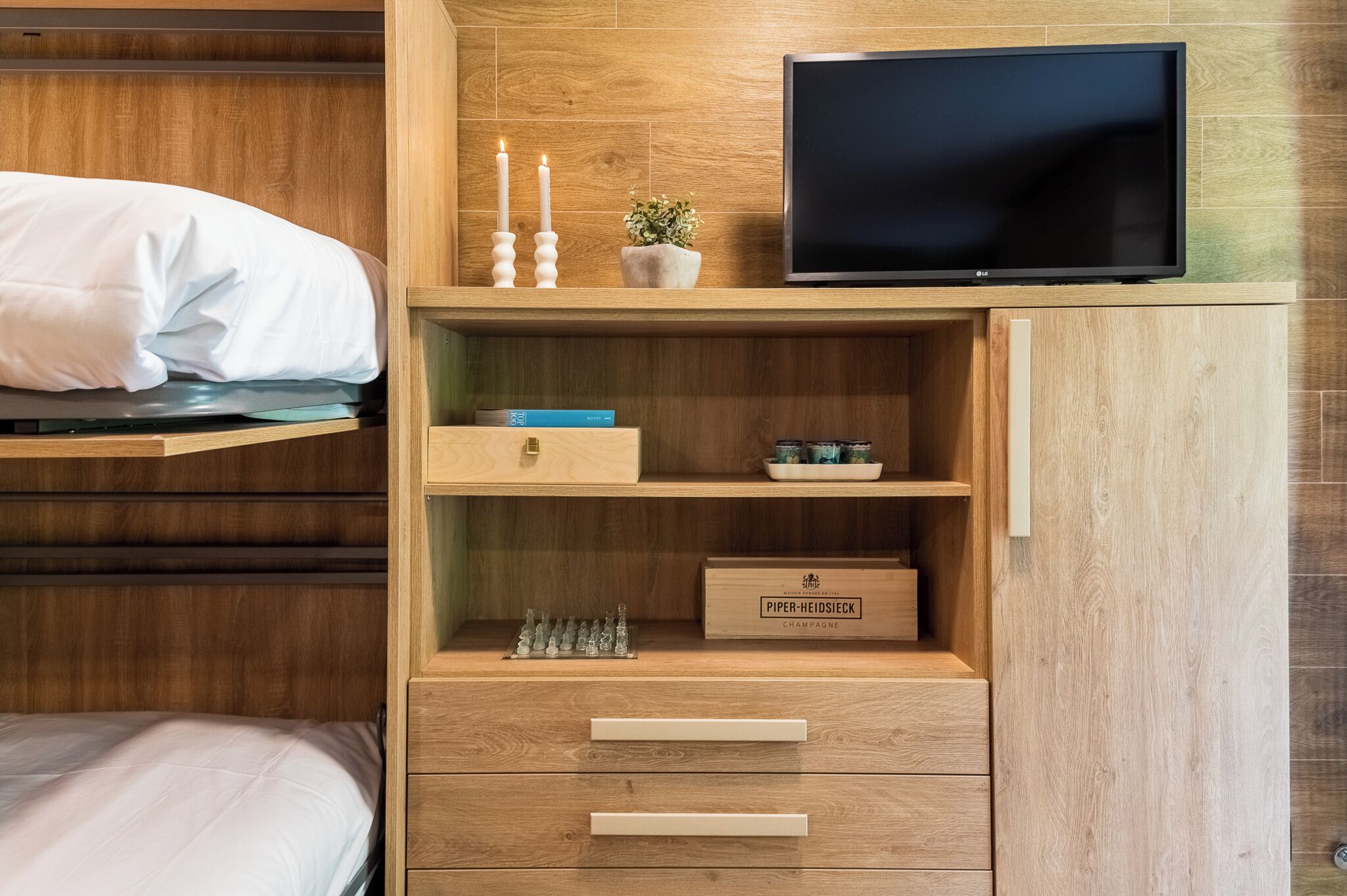 A bedroom with a bed, a TV, wooden shelves, a small plant, and a champagne box.