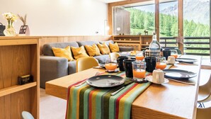 Ein moderner Essbereich mit einem Holztisch, gedeckt für ein Frühstück, einer Couch mit gelben Kissen und Blick auf Berge durch große Fenster.
