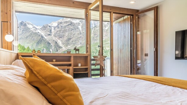 Ein Schlafzimmer mit einem Bett, einem gelben Kissen, einem Nachttisch aus Holz, einem Regal mit Dekoration und Blick auf Berge durch große Fenster.