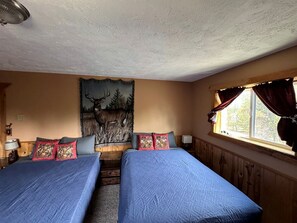 A room with two beds, a tapestry with a deer, a window with curtains, and a wooden wall panel. - Incredible Cabin Perfect for Family Reunions near Yellowstone, Idaho (Island Park)