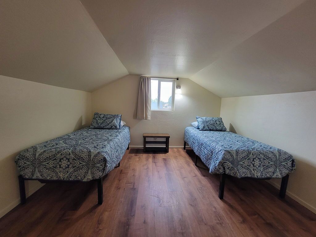 Habitació petita, amb un llit individual per a dues persones, sòl de fusta, finestra amb cortina i una taula petita.