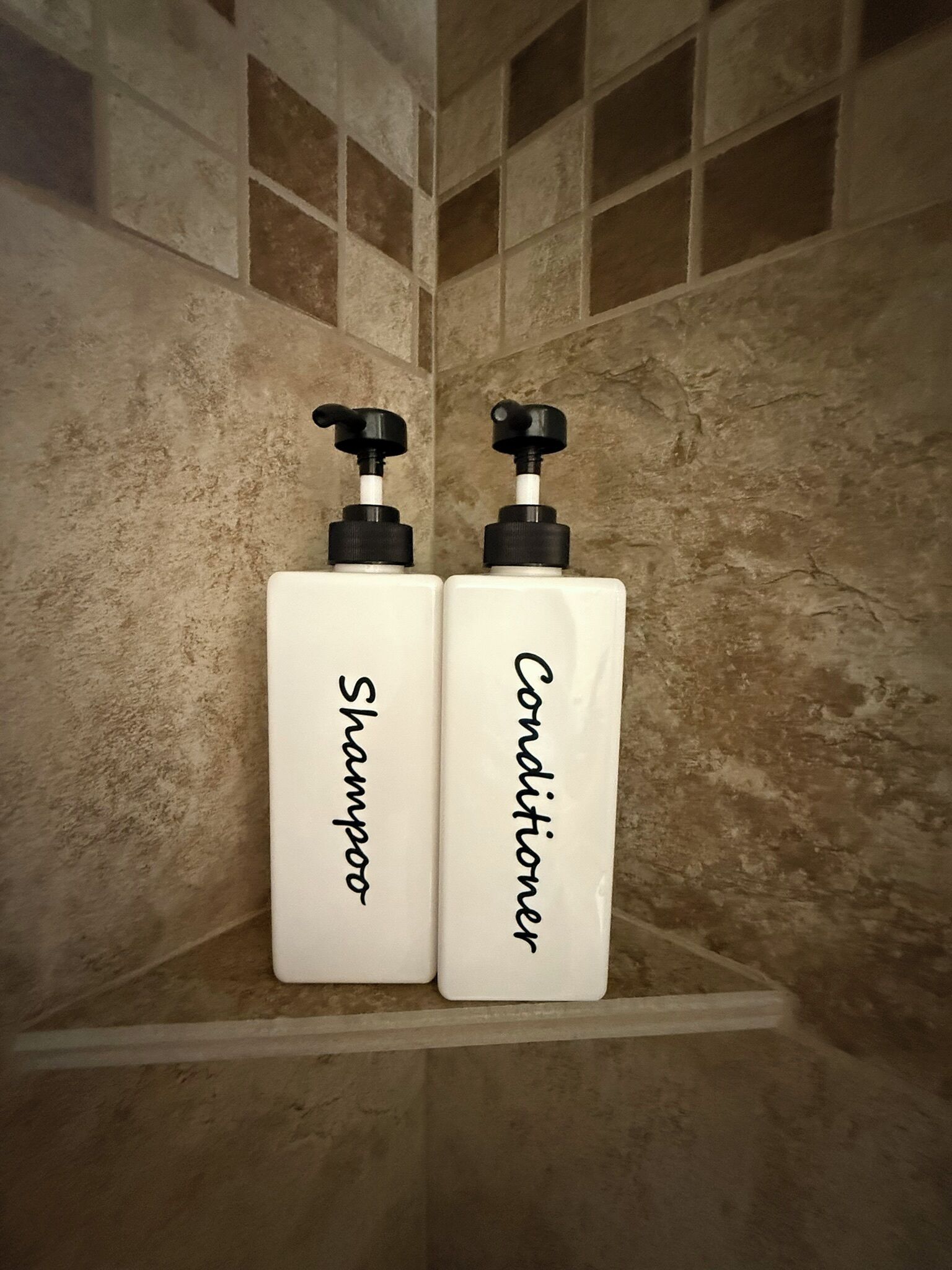 Two white bottles labeled 'Shampoo' and 'Conditioner' on a shelf in a bathroom with tiled walls.