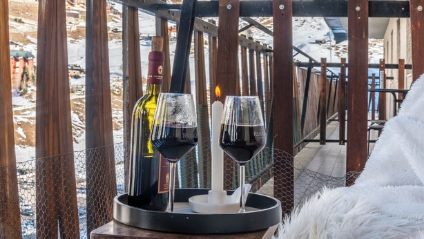 Un tavolo con due bicchieri di vino rosso, una candela e una bottiglia di vino, sullo sfondo di un paesaggio innevato con una ringhiera in legno.