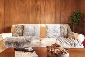 A living room with a wooden panel wall, a white sofa with multiple cushions, a remote control, a book, and a decorative vase.