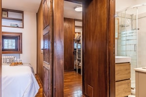 A hotel room with a wooden wardrobe, a bed with a tray of bread, a white bathroom with a glass shower enclosure, and a view of the outdoors.