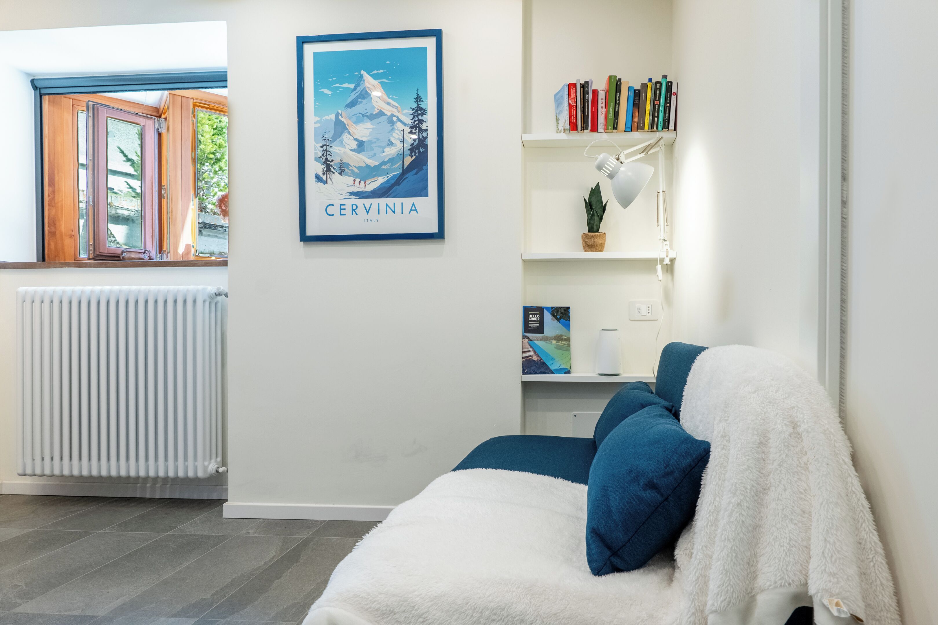 Een slaapkamer met een bed, een radiator, een raam met uitzicht op een berg, een boekenkast met boeken en een lijst aan de muur.