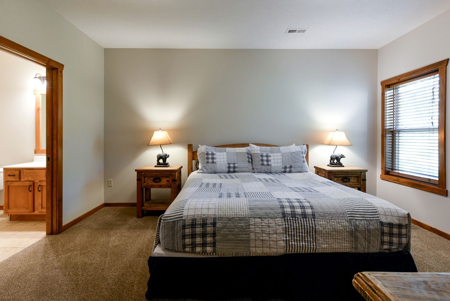 A bedroom with a large bed, two bedside tables with lamps, a wooden headboard, a window with blinds, and a door leading to another room.