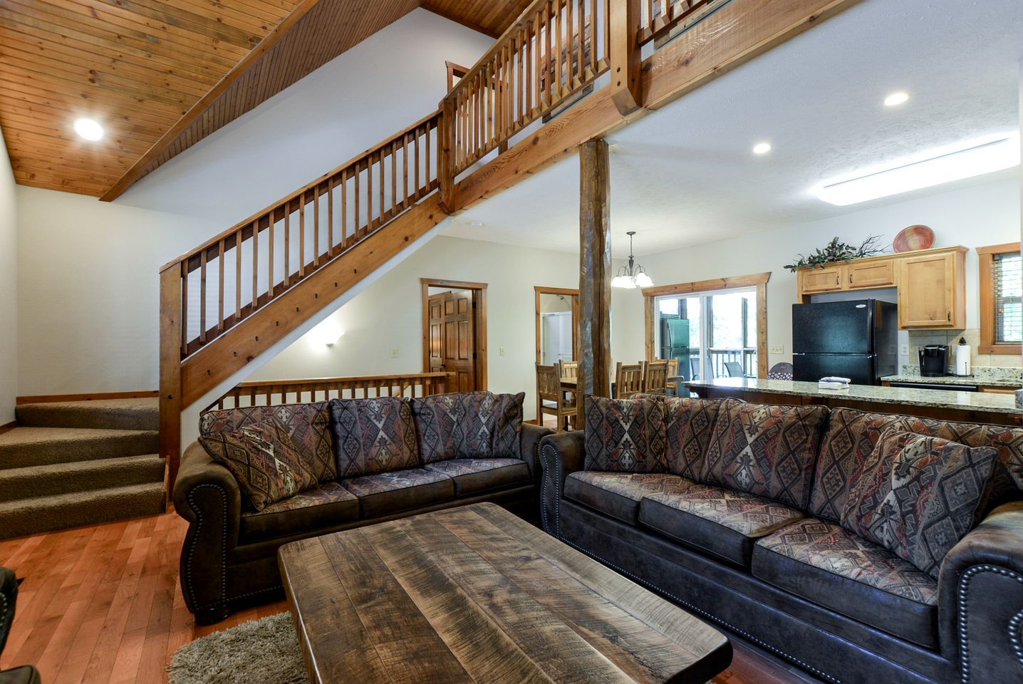 A spacious living room with a wooden staircase, a large sofa set, and a kitchen area with wooden cabinets and a refrigerator.