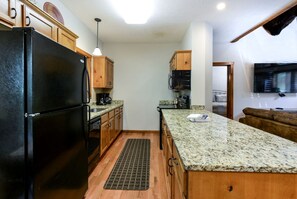 Eine Küche mit einem schwarzen Kühlschrank, Holzschränken, Granitarbeitsplatten, einer Mikrowelle und einem Fernseher im angrenzenden Wohnbereich.