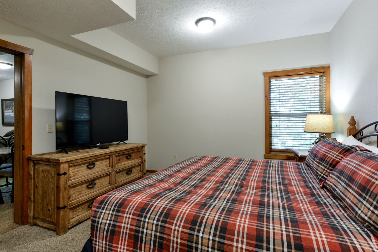 A bedroom with a plaid bedspread, a wooden dresser, a television on top, and a window with blinds.