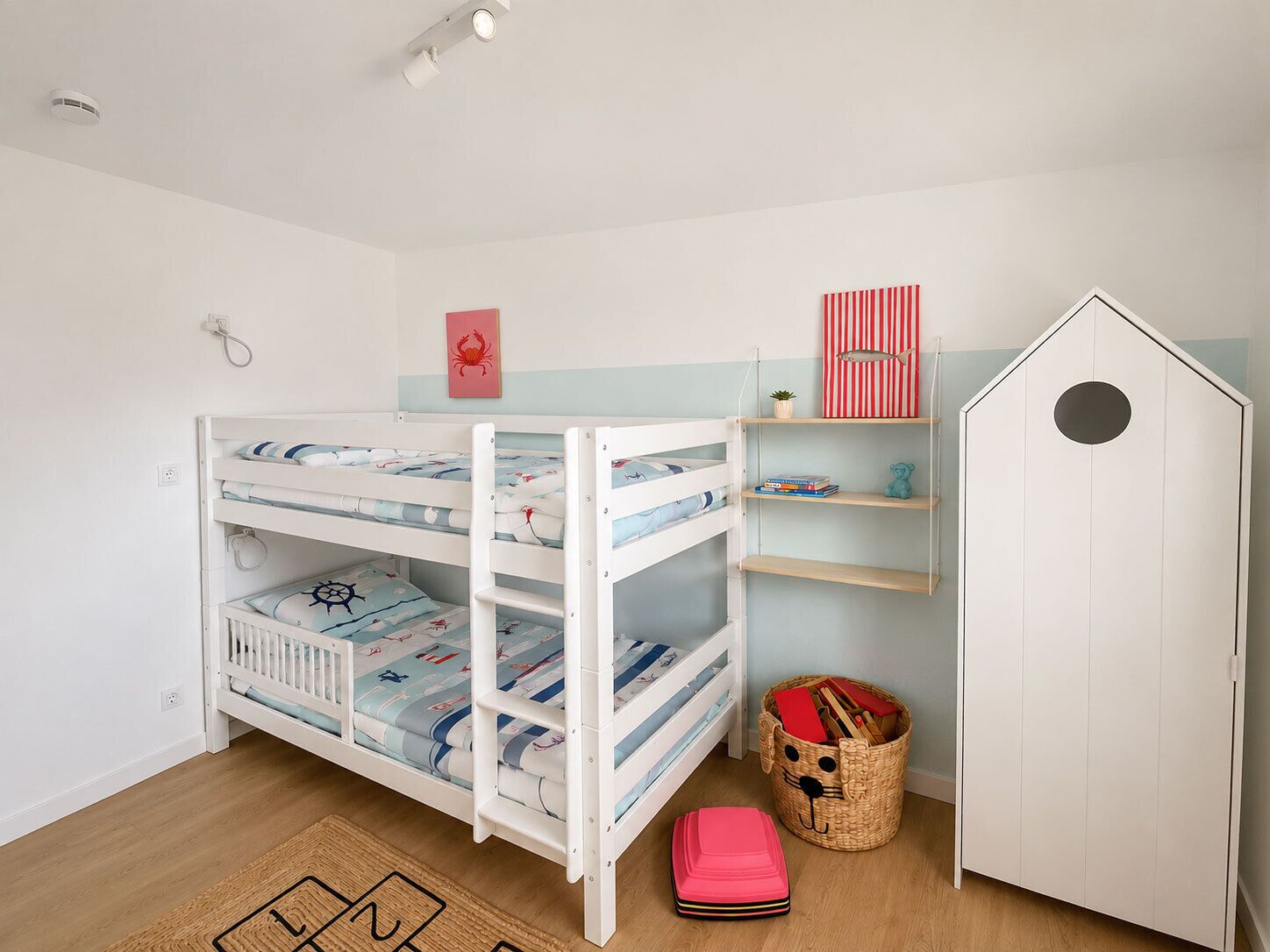 Ein Stockbett mit Meeresmotiven, ein kleines Regal mit Büchern und Spielzeug, ein Korb voller Spielsachen und ein weißer Schrank mit runder Aussparung.