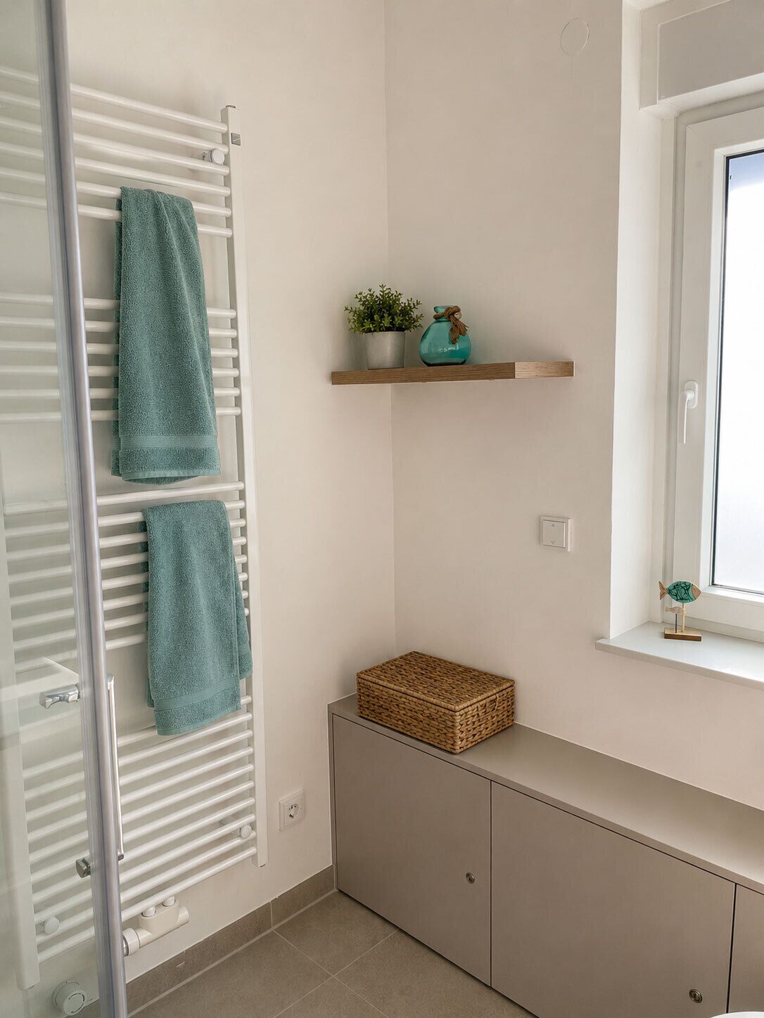 Une salle de bain avec un porte-serviettes, une étagère ornée d’une plante et d’un objet décoratif, une boîte en osier et une fenêtre.
