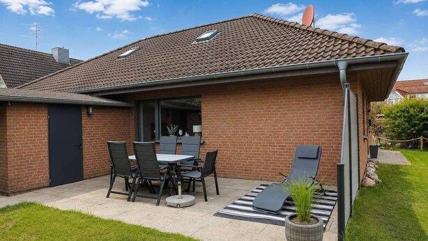 Een huis van baksteen met pannendak, een terras met tafel en stoelen, en een kleine tuin.