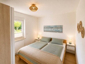 A bedroom with a bed, two bedside tables, a window, a wall-mounted light fixture, and a decorative sign. - Kellenquartier – Anker 8 | Newly renovated, family-friendly, with a garden (Kellenhusen (Ostsee))