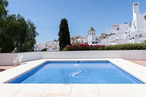 Ein Swimmingpool mit klarem, blauem Wasser, umgeben von weißen Gebäuden und einem weißen Zaun. Ein einzelner weißer Stuhl steht daneben.