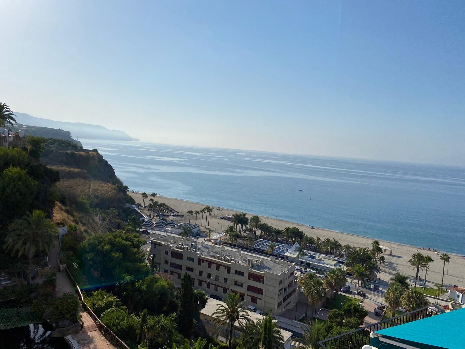 A coastal view with a beach, palm trees, and a building with multiple floors and balconies.