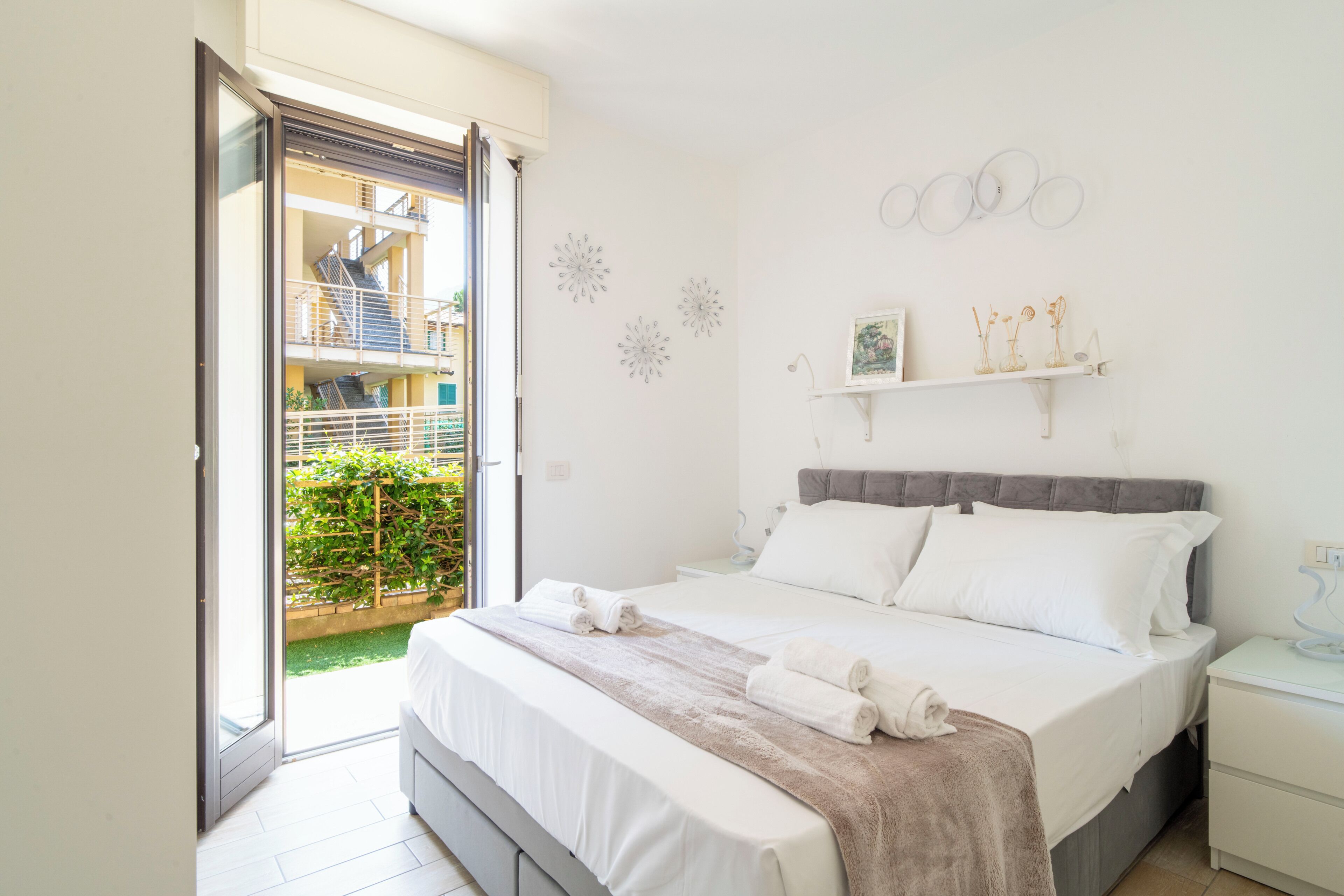 Een slaapkamer met een bed, nachtkastje, raam en uitzicht op een balkon.