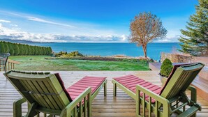 En terrass med två gröna Adirondack-stolar och en röd kudde, med utsikt över en gräsyta och havet.