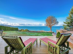 A deck with two green Adirondack chairs and a red cushion, overlooking a grassy area and the ocean. - Waterfront Retreat: Hot Tub, Sauna & Beach Access (Bellingham)