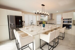 A modern kitchen with a central island, white bar stools, stainless steel appliances, and a pendant light fixture. - Horizon- 6BR Gated Villa Near Beach w Pool (Oistins)