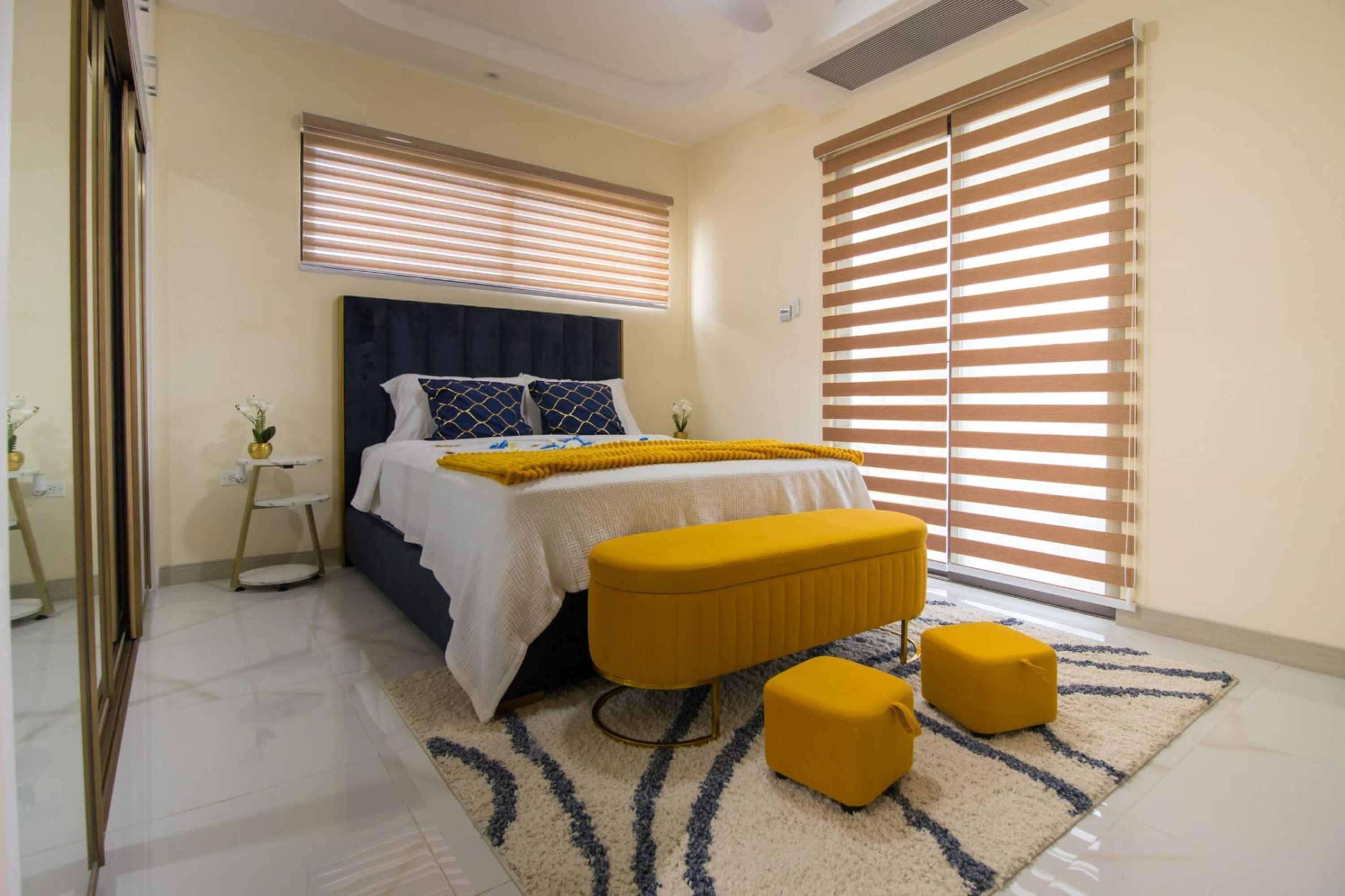 A bedroom with a bed, bedside tables, a bench, and large windows with blinds.