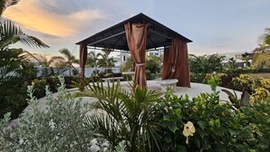 A covered outdoor seating area with a table and chairs, surrounded by lush greenery and a clear sky. - Aurelia- 1BR Luxury Beachfront Condo (St. Ann's Bay)