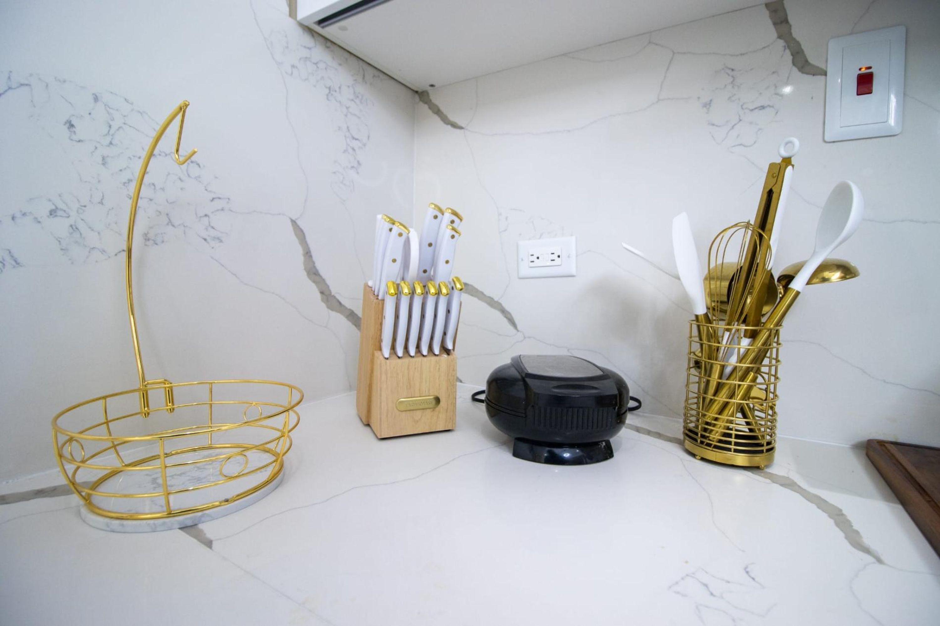 A kitchen with a marble countertop, a gold wire fruit basket, a knife block, a black appliance, and a gold utensil holder.