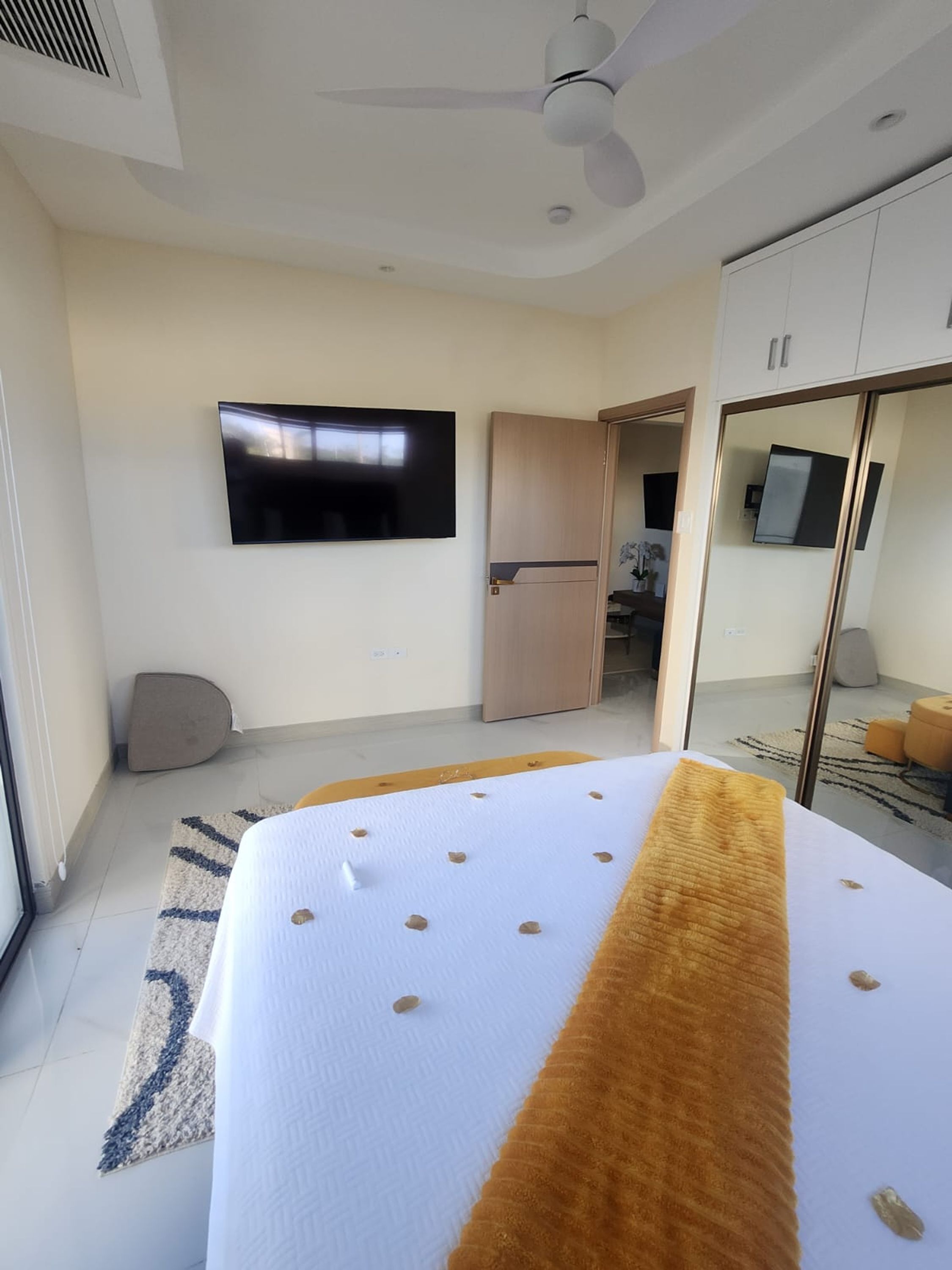 A hotel room with a bed, a flat-screen TV mounted on the wall, a ceiling fan, and a wardrobe with a mirror.