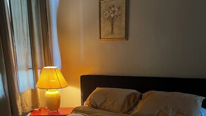 Ein ordentlich bezogenes Bett mit weißer Daunendecke, ein Nachttisch mit Lampe und ein gerahmtes Kunstwerk an der Wand.
