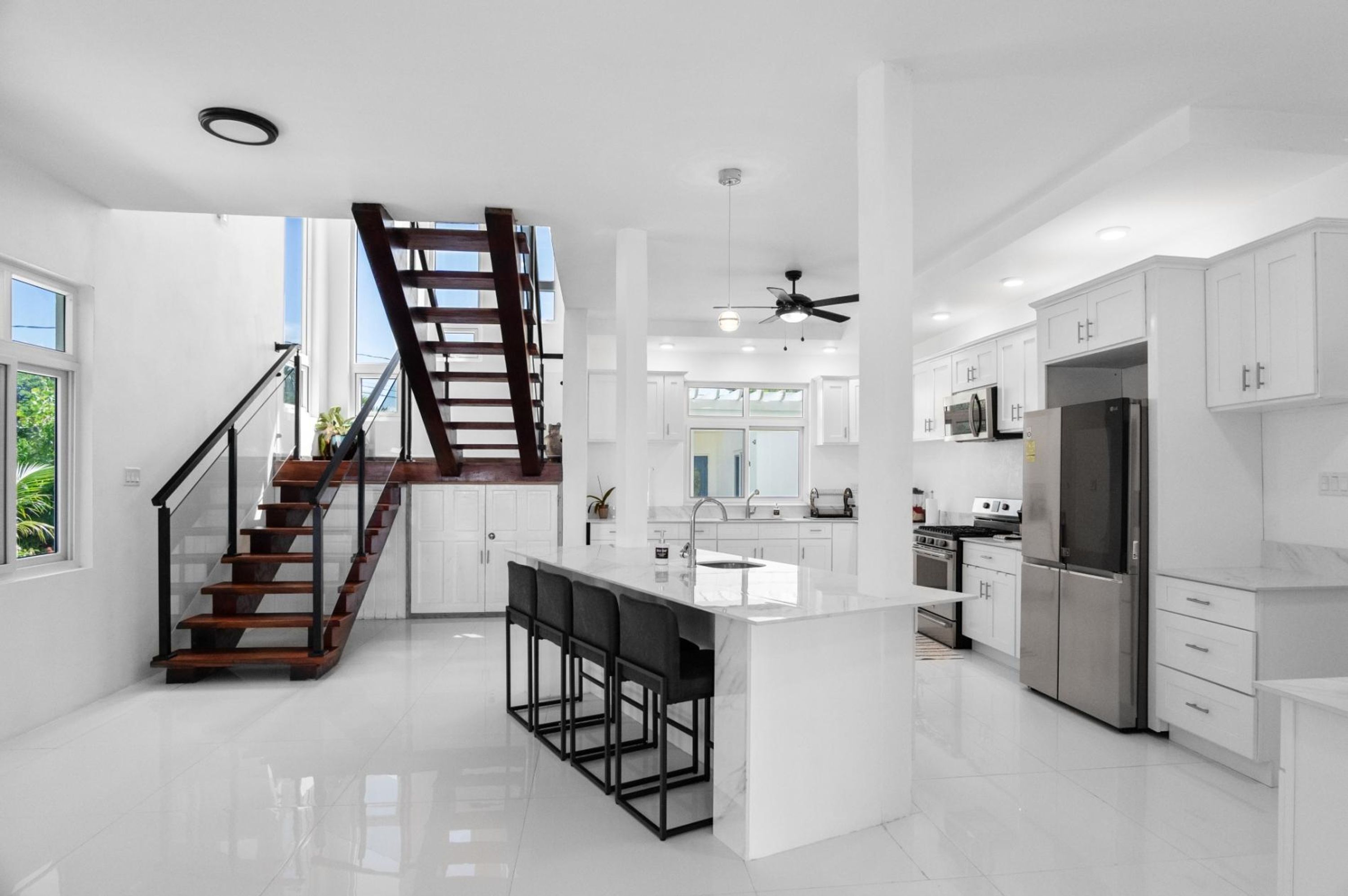 A modern kitchen with white cabinetry, a central island with bar stools, a stainless steel refrigerator, and a wooden staircase.