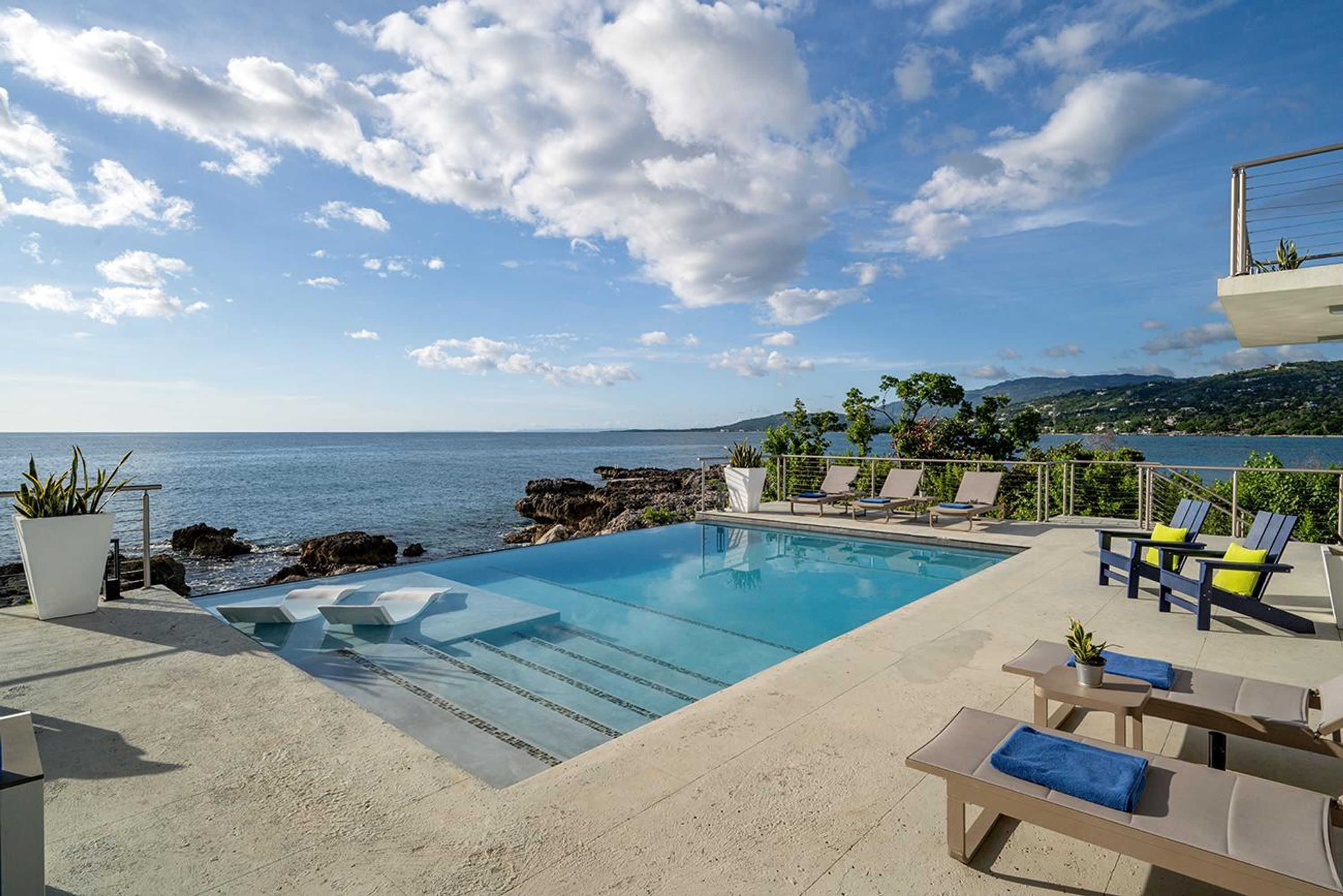 A poolside area with lounge chairs, a pool, and a view of the sea.