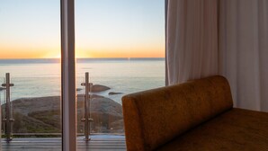 Een kamer met een groot raam met uitzicht op de kust bij zonsondergang, een houten terras en een bruine bank.