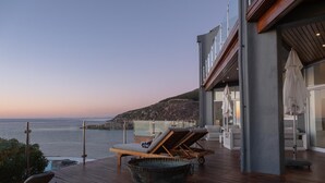 Een houten terras met lounge stoelen, een grote schaal, en uitzicht op zee en een rotsachtige heuvel.