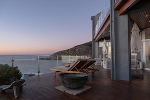 Eine Holzterrasse mit Liegestühlen, einer großen Schüssel und Blick auf das Meer und einen felsigen Hügel.