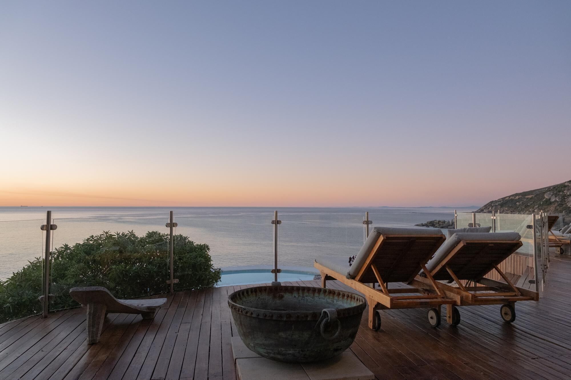 A wooden deck with lounge chairs, a large bowl, and a view of the sea at sunset.