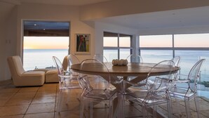 Een moderne eetruimte met een ronde tafel en transparante stoelen, grote ramen met uitzicht op zee, en een beige bank.