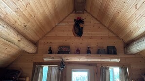 A wooden cabin interior with a sloped ceiling, a bed with patterned bedding, two lamps, a door with a view of a snowy landscape, and a shelf with various items.