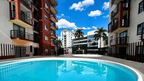 A swimming pool surrounded by multi-story buildings with balconies, a clear sky, and palm trees. - Apartamento en Pinares con piscina (Pereira)