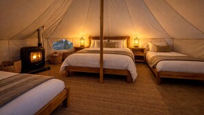 A tent with two beds, a wood-burning stove, and a view of trees through a window.