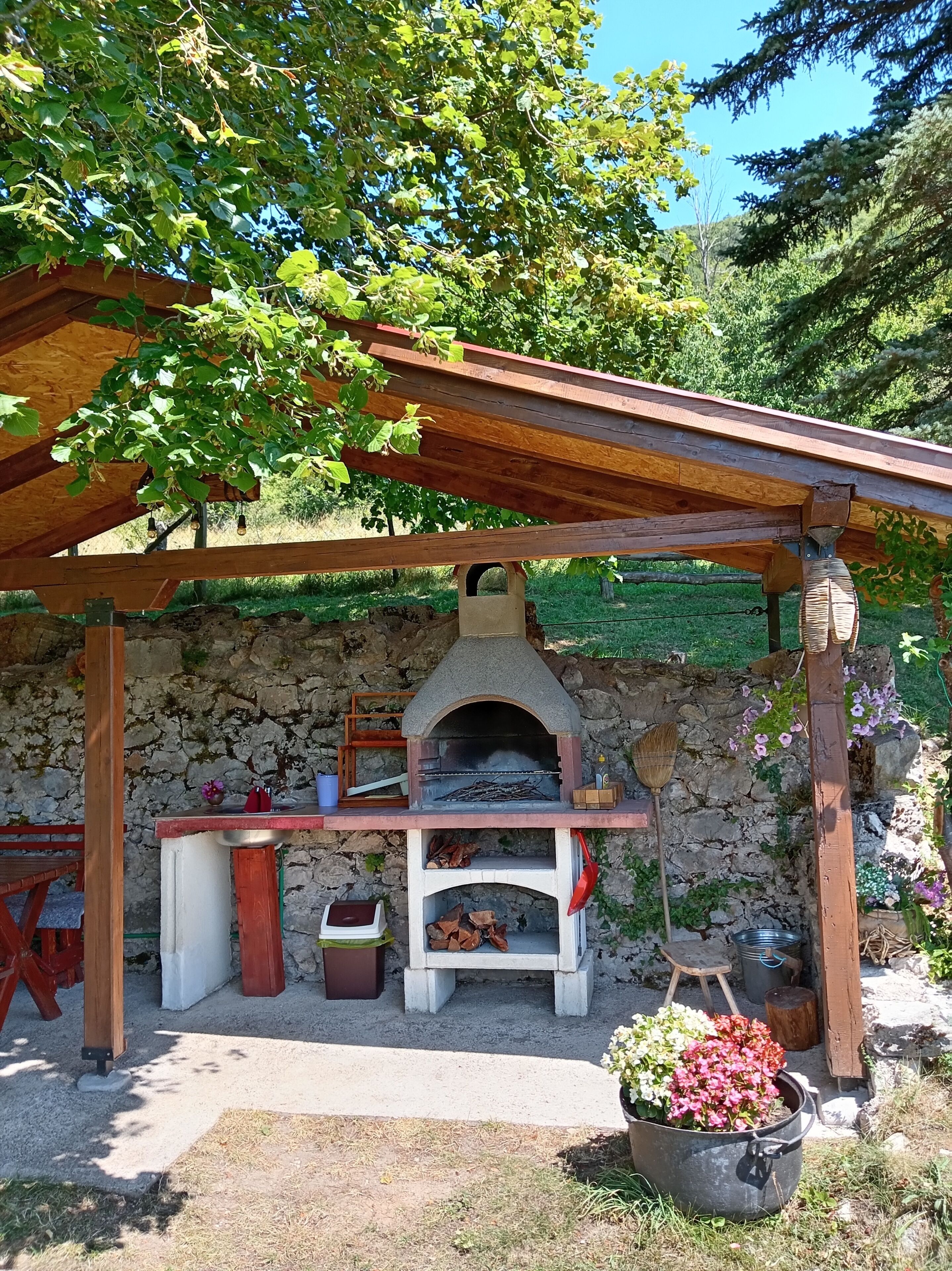 An outdoor stone oven with a wooden structure overhead, a fire pit, and a rustic setting with greenery.