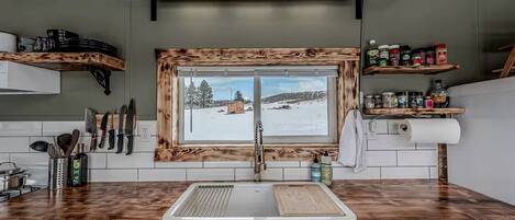 A kitchen with a wooden countertop, a window with a snowy view, and a shelf with kitchen utensils.