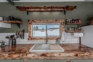 Une cuisine avec un plan de travail en bois, une fenêtre donnant sur un paysage enneigé et une étagère remplie d’ustensiles de cuisine.
