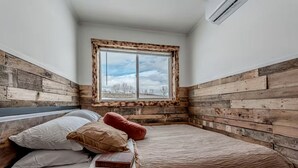 A bedroom with a wooden headboard, a window with a view, and a bed with pillows and a book. - Serene Recycled Container in Big Sky Montana, Perfect for a Glamping Weekend in the Wilderness (Ovando)