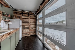 A kitchen with a wooden accent wall, a white refrigerator, and a window with a snowy view. - Serene Recycled Container in Big Sky Montana, Perfect for a Glamping Weekend in the Wilderness (Ovando)